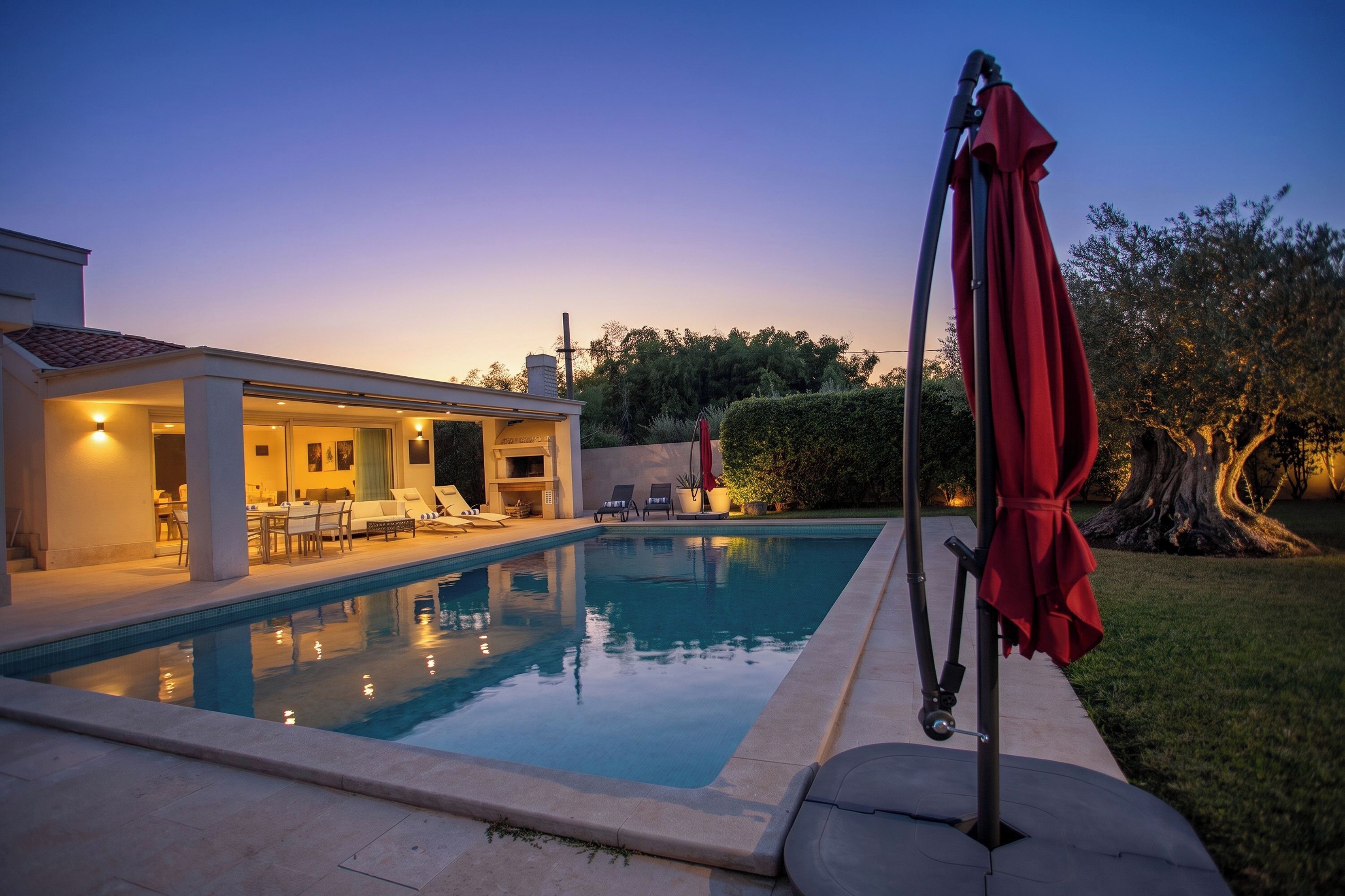 Una piscina con sillones de descanso y un área cubierta, rodeada de un césped cuidado y árboles.