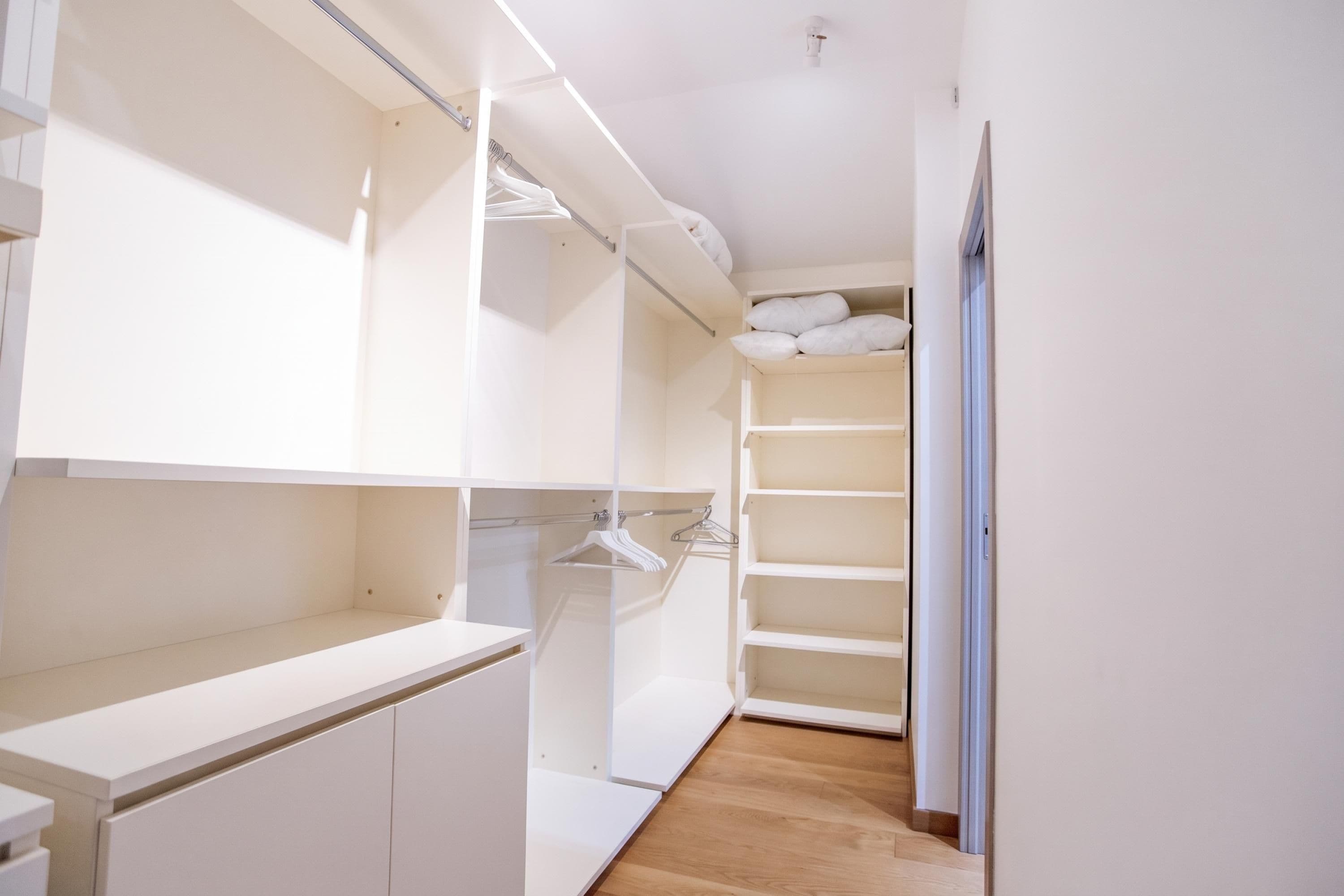 Un vestidor estrecho con estantes blancos y espacio para colgar, piso de madera y una puerta blanca.