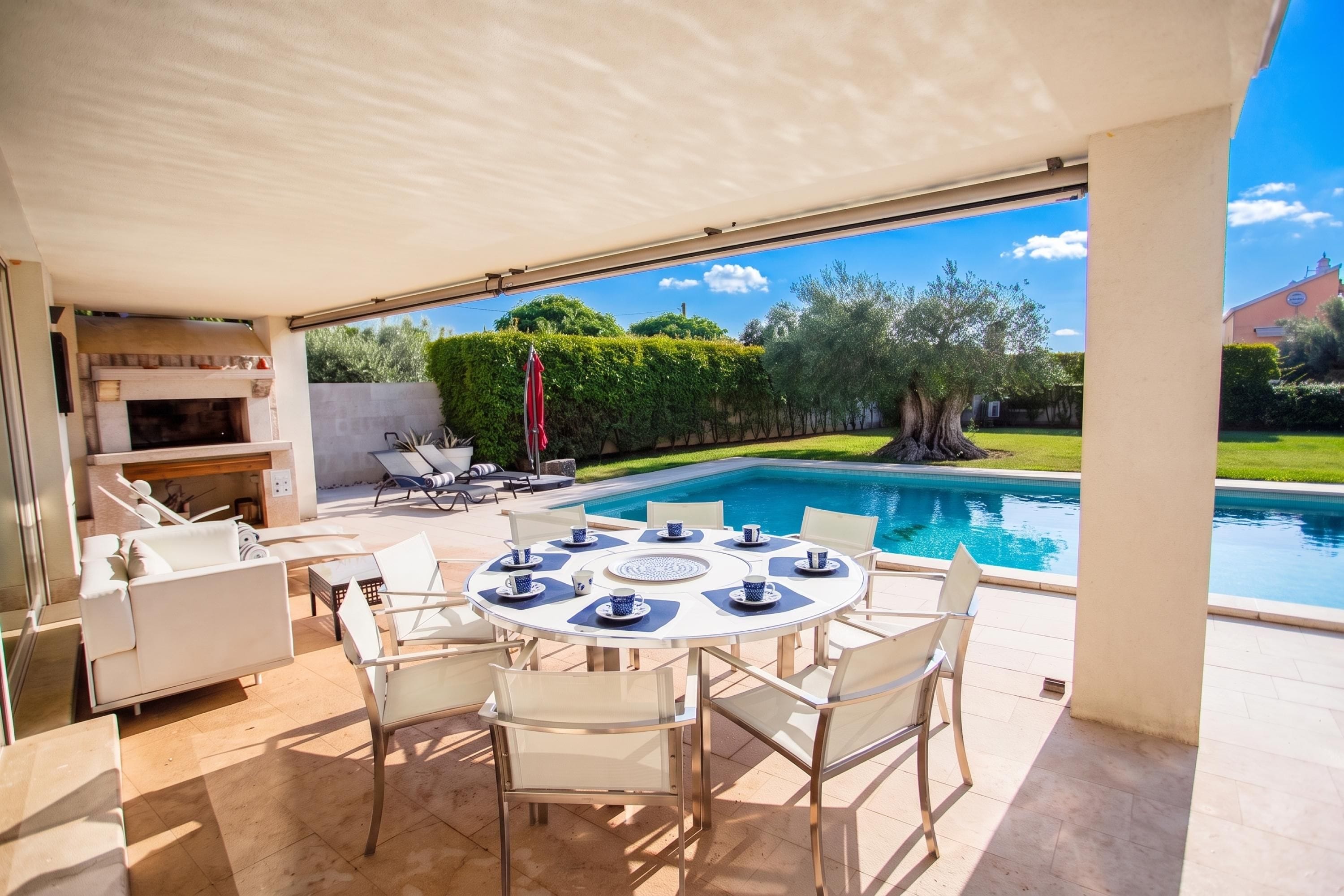 Un espace repas extérieur moderne, aménagé à proximité d’une piscine, comprenant une table ronde avec des chaises, un barbecue et un parasol.