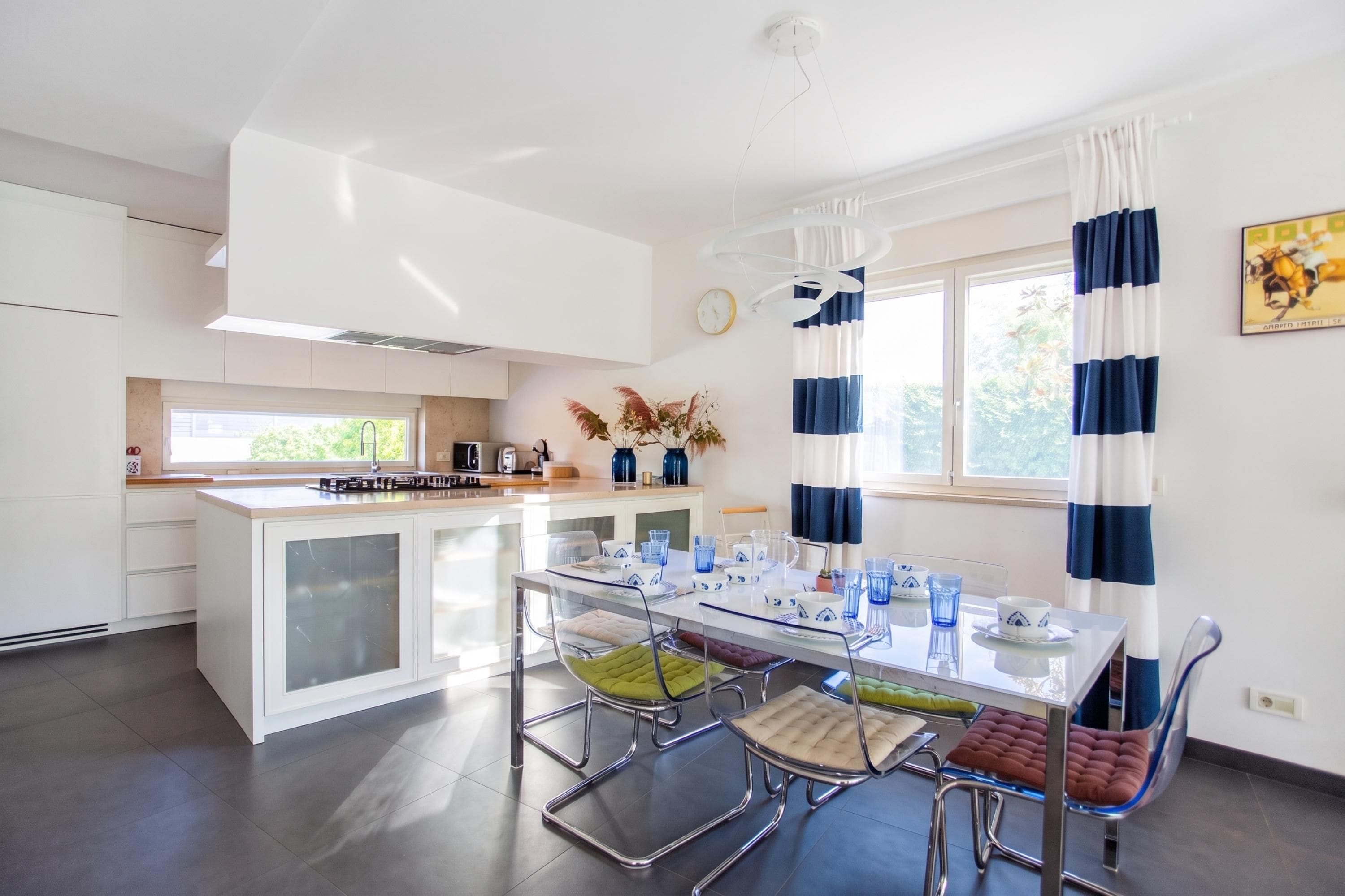 A modern kitchen with a central island, a dining area with a glass table, and a view of the outdoors.