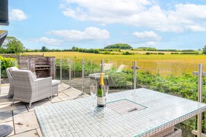 Een terras buiten met rieten meubels, een glazen tafel en een fles champagne.
