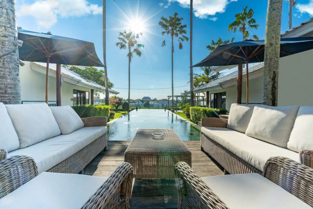 A pool area with lounge chairs and umbrellas, surrounded by palm trees and a clear sky.