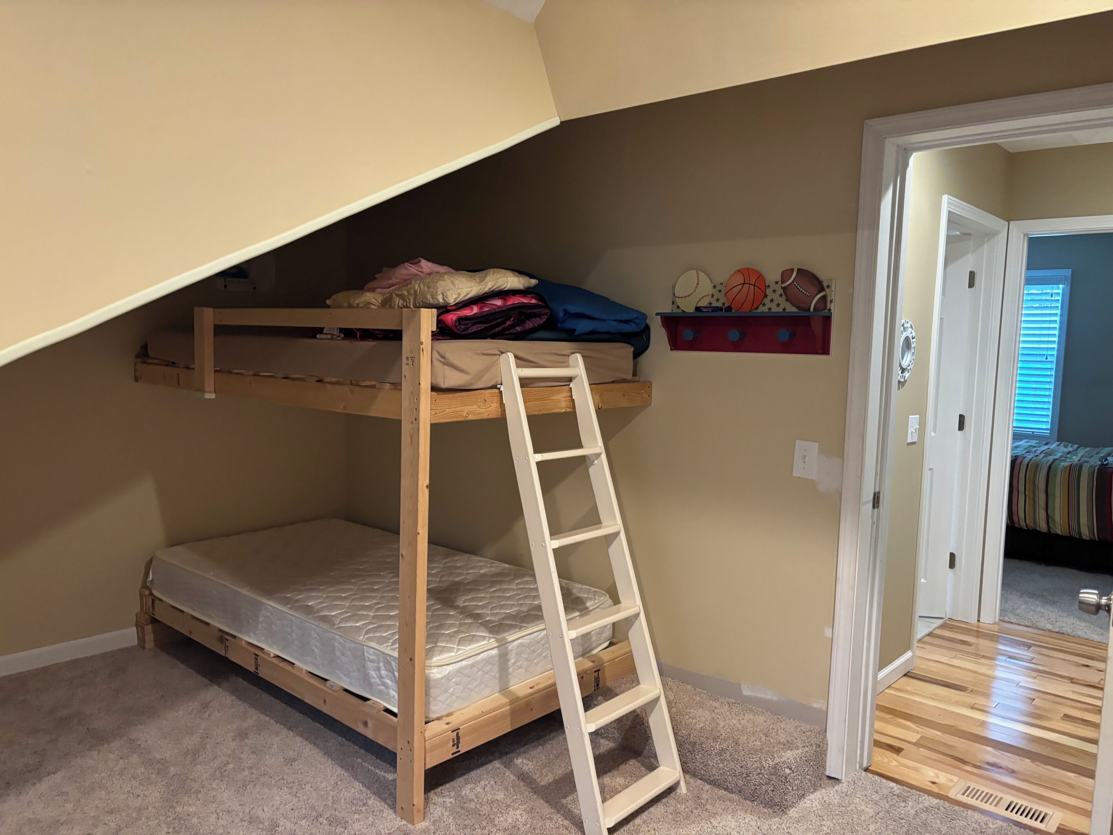Een stapelbed met een ladder, een plank met sportdecoraties, en een deur naar een andere kamer.