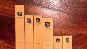 Un set di sei scatole di cartone con il logo dell''AKSAI Boutique Hotel', etichettate come 'KIT IGIENE DENTALE', 'PETGNE', 'KIT RASOIO', 'SAPONE' e 'CAPPELLO DA DOCCIA'.
