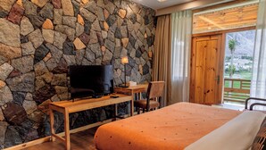Una camera da letto con parete in pietra, una TV su una scrivania in legno, un letto con coperta arancione e vista su un paesaggio montuoso attraverso una porta in legno.