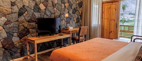 A bedroom with a stone wall, a TV on a wooden desk, a bed with an orange blanket, and a view of a mountainous landscape through a wooden door.