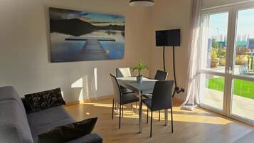 Ein modernes Wohnzimmer mit Essbereich, einem großen Fenster mit Vorhängen, einem Flachbildfernseher und einem Gemälde einer Landschaft mit See.
