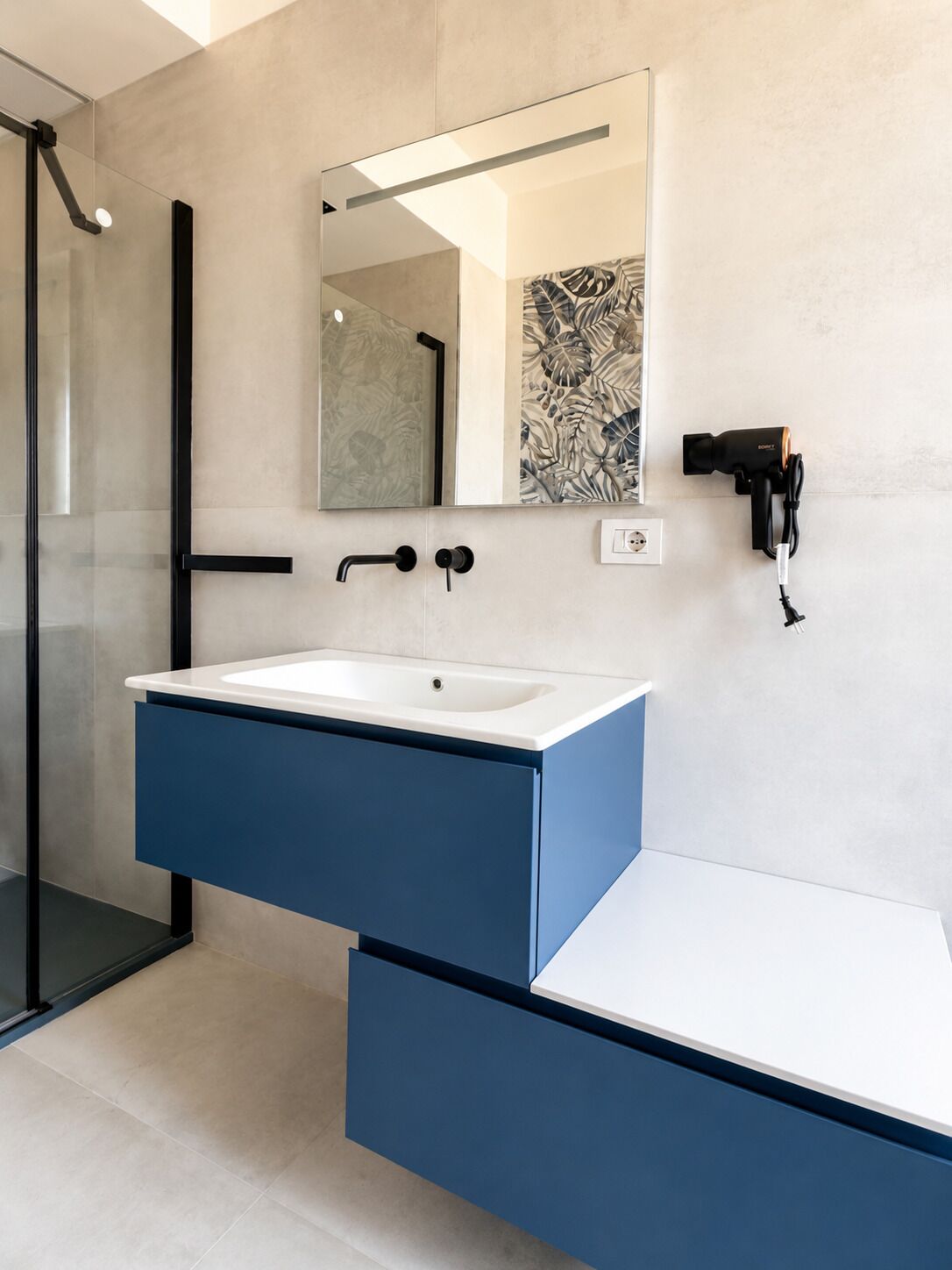 Un baño moderno con lavamanos blanco, tocador azul, cabina de ducha de vidrio y un secador de pelo montado en la pared.