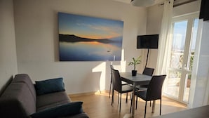 A living room with a sofa, a dining table with chairs, a painting on the wall, and a TV mounted on the wall. - Seven Lakes ApartHotel RTA (Burolo)
