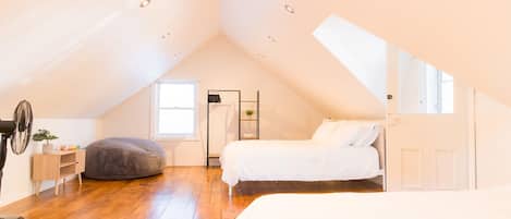 Un amplio dormitorio en ático con techo inclinado, suelo de madera, un ventanal grande y una cama con ropa de cama blanca.