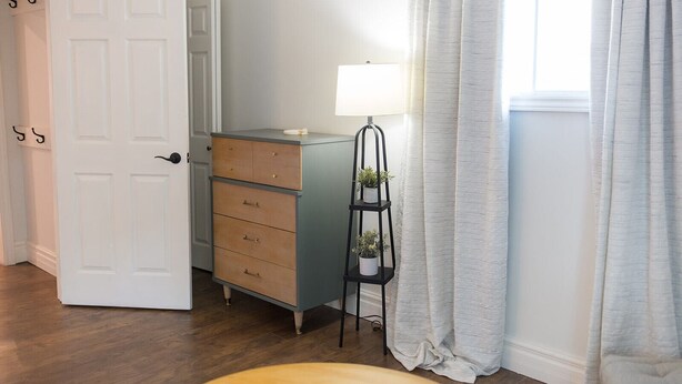 Une pièce avec un plancher en bois, une porte blanche, une commode bleue avec des tiroirs, une petite table avec une lampe et des plantes, et une fenêtre avec des rideaux.