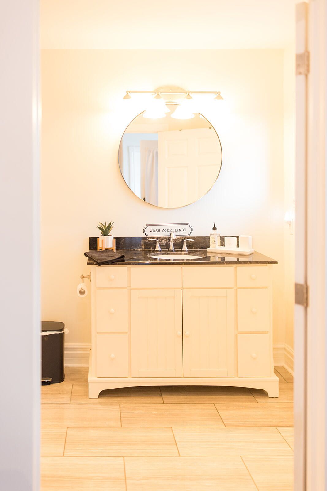A bathroom with a vanity, a round mirror, and a 'WASH YOUR HANDS' sign.