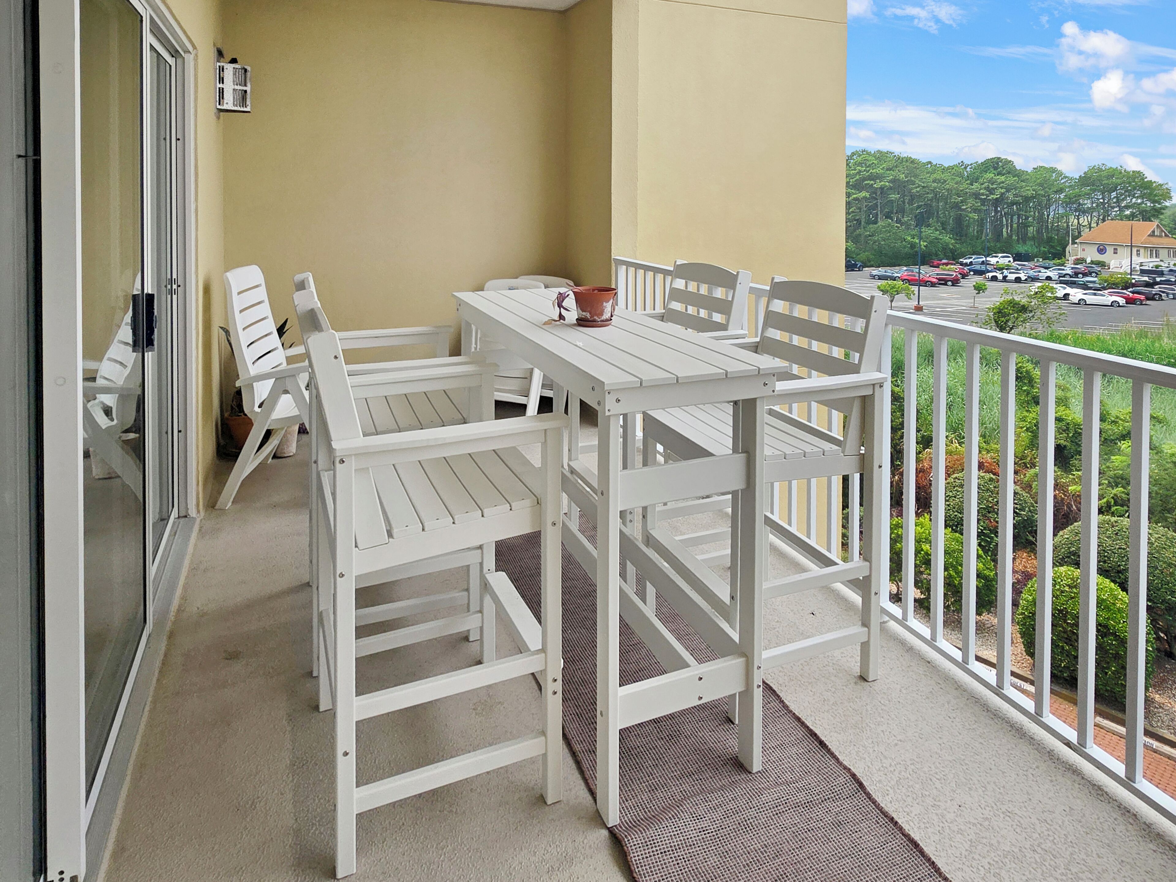 Un balcon avec une table et des chaises blanches, un pot de fleurs et une vue sur un parking et des arbres.