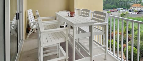 Un balcon avec une table et des chaises blanches, un pot de fleurs et une vue sur un parking et des arbres.