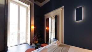 A hotel room with a bed, a small table, and a chair. There are two windows with curtains, and a view of the outside. - Sancta Nolana (Naples)