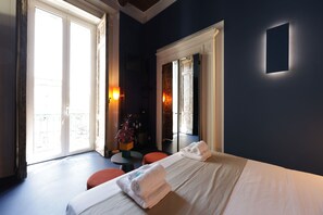 Quarto de hotel com cama, uma pequena mesa e uma cadeira. Há duas janelas com cortinas, e vista para o exterior.