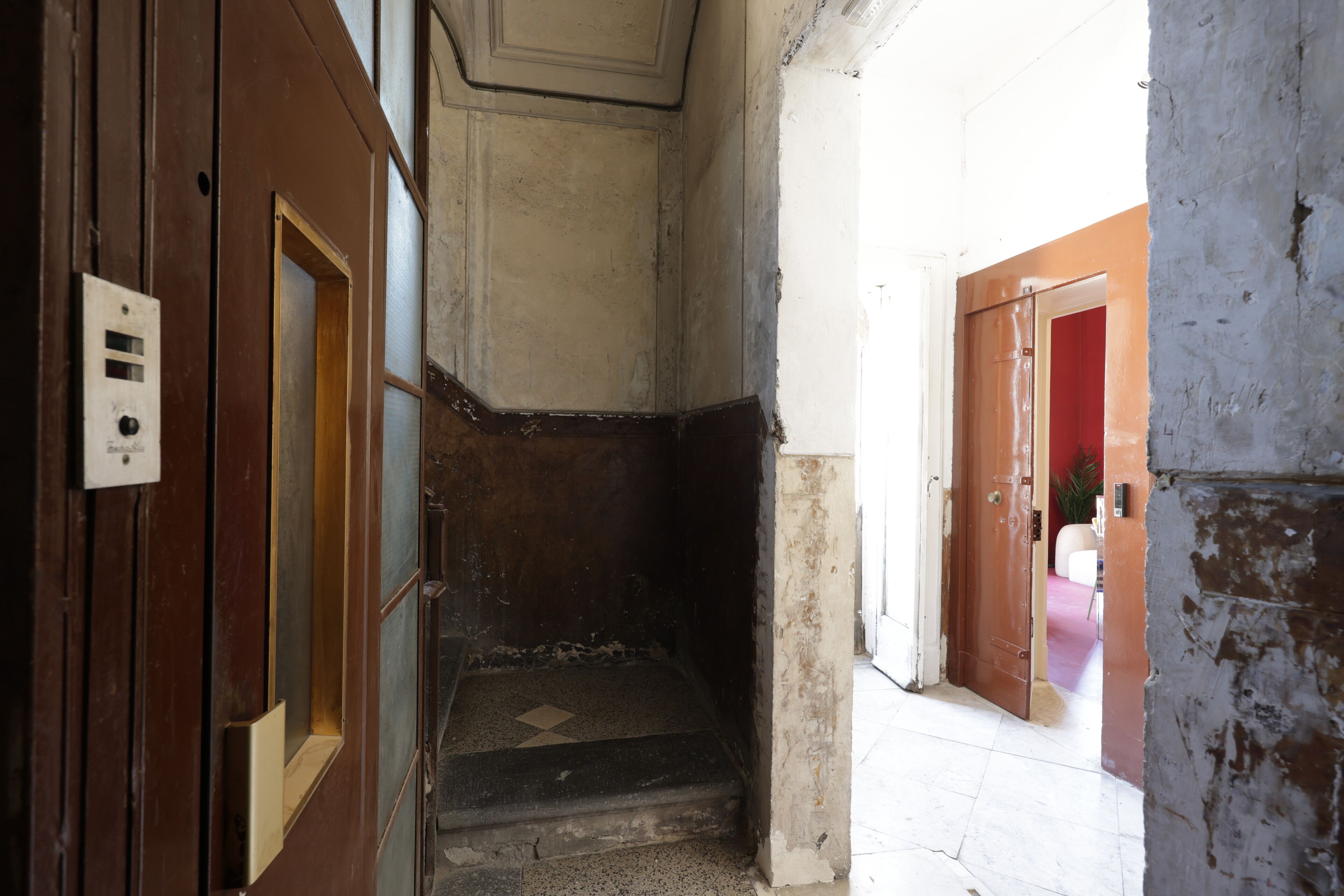 Eine schmale Treppe, die zu einer Zwischenebene führt, mit einer Tür links und einer roten Tür rechts.