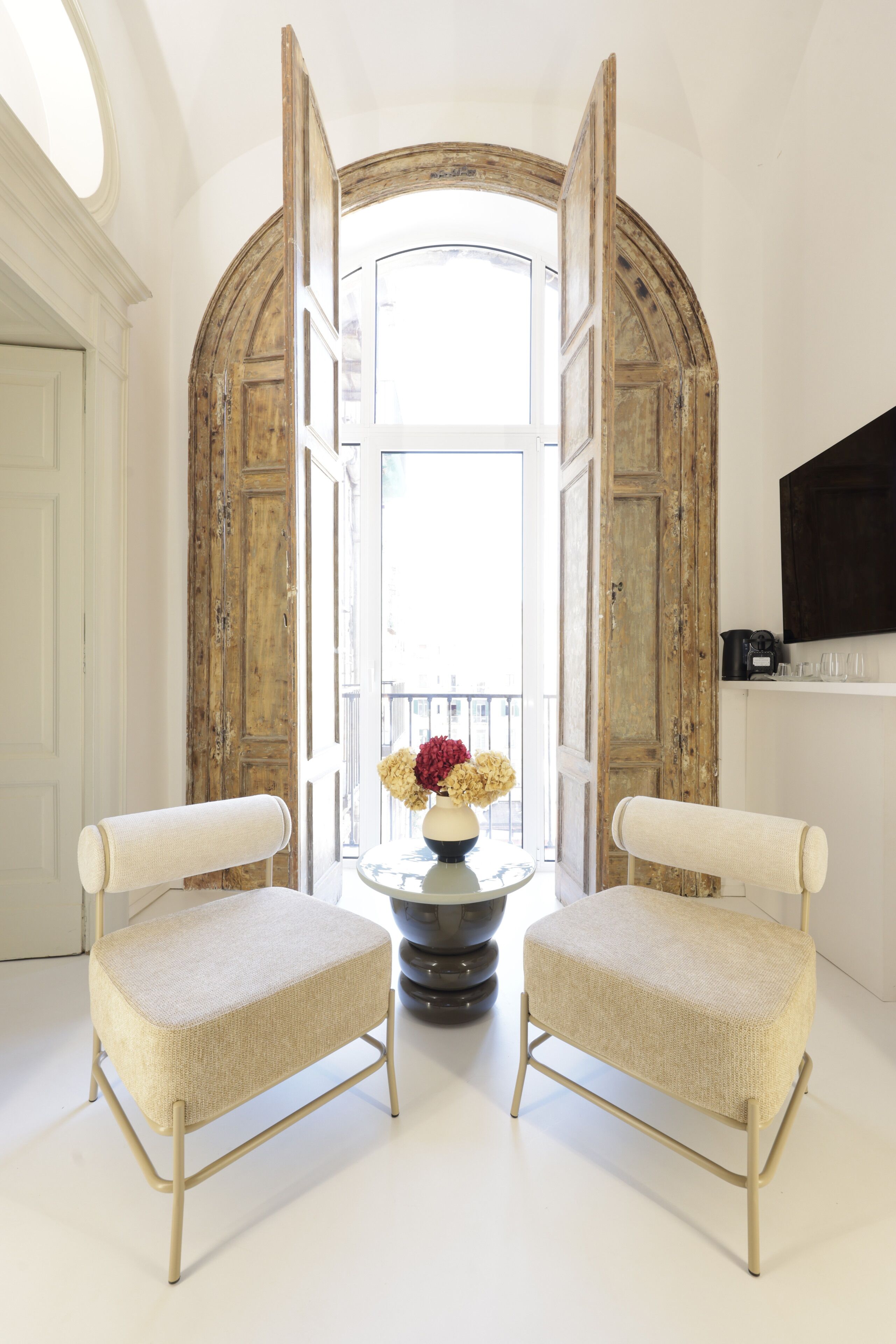 A room with two upholstered chairs, a round table with a vase of flowers, and a large arched doorway.