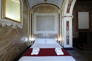 Quarto de hotel com uma cama grande, colcha vermelha e parede de mármore.