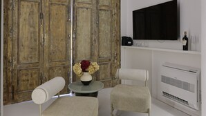 A room with a large wooden door, a television on a white shelf, a small table with a vase, and two upholstered chairs. - Sancta Nolana (Naples)