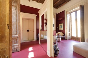 A spacious room with a red floor, white walls, and a wooden ceiling. There is a large white door, a chair, and a table with a vase of flowers. - Sancta Nolana (Naples)