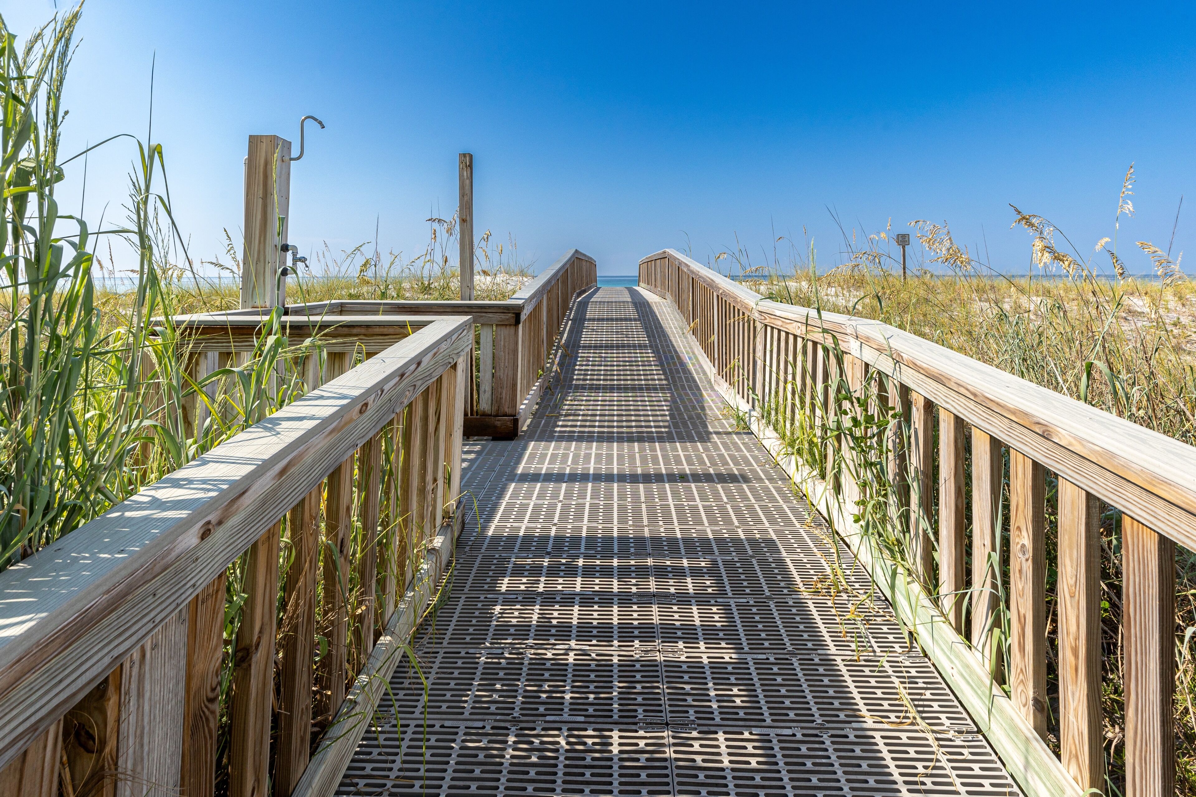 A wooden boardwalk leading to a beach, surrounded by tall grass and clear blue sky.