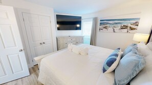 A hotel room with a large bed, a flat-screen TV, a painting of a beach scene, and a lamp. - Islander Beach Resort 506 2 Bedroom Condo (Fort Walton Beach)
