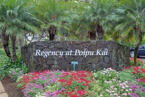 Eine Steinsäule mit dem Schriftzug 'Regency at Poipu Kai', umgeben von bunten Blumen und Palmen.
