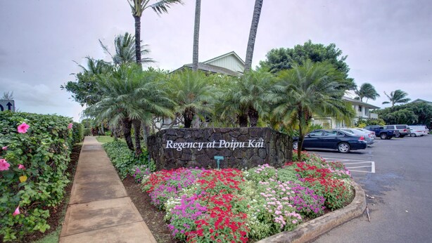 A stone wall with 'Regency at Poipu Kai' signage, surrounded by tropical plants and a parking area with cars. - Kauai Regency At Poipu Kai #823 3 Bedroom Condo (Koloa)