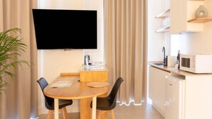 Une chambre moderne dotée d’un téléviseur à écran plat fixé au mur, d’une table à manger en bois avec des chaises noires, d’un coin cuisine équipé d’un four à micro-ondes et d’une plante.