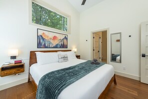 A bedroom with a bed, a nightstand, a bedside table, a wall-mounted painting, a mirror, and a door leading to another room. - Murray Creek Hollow - Private Asheville Escape (Asheville)