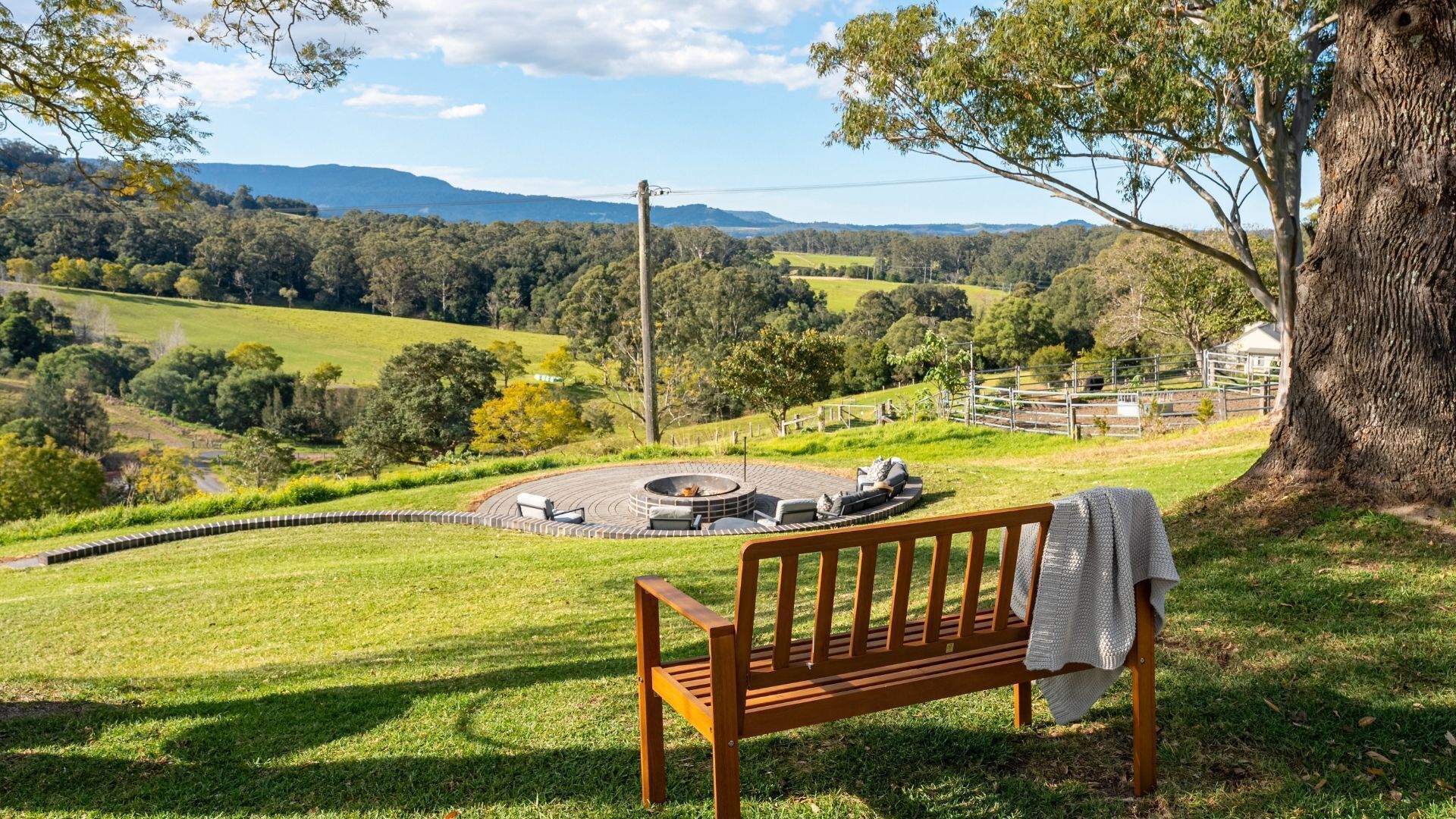 A wooden bench overlooking a circular outdoor seating area with a fire pit, set against a backdrop of a lush green landscape and distant hills.