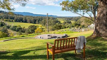 A wooden bench overlooking a circular outdoor seating area with a fire pit, set against a backdrop of a lush green landscape and distant hills.