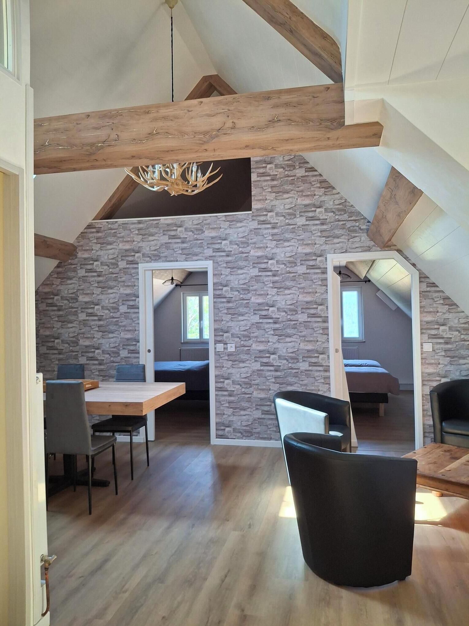 A spacious loft with exposed wooden beams, a stone wall, a dining area with a table and chairs, and a black leather armchair.