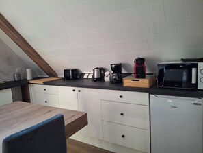 Eine kleine Küche mit weißen Schränken, einem Holztisch und verschiedenen Geräten, darunter eine Kaffeemaschine, ein Wasserkocher und eine Mikrowelle.