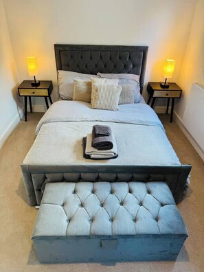 A bedroom with a tufted headboard, a bed with a light-colored duvet and pillows, a bedside table with a lamp, and a matching ottoman. - #13 Elegant Solihull Townhouse Sleeps 10 NEC/BHX (Solihull)