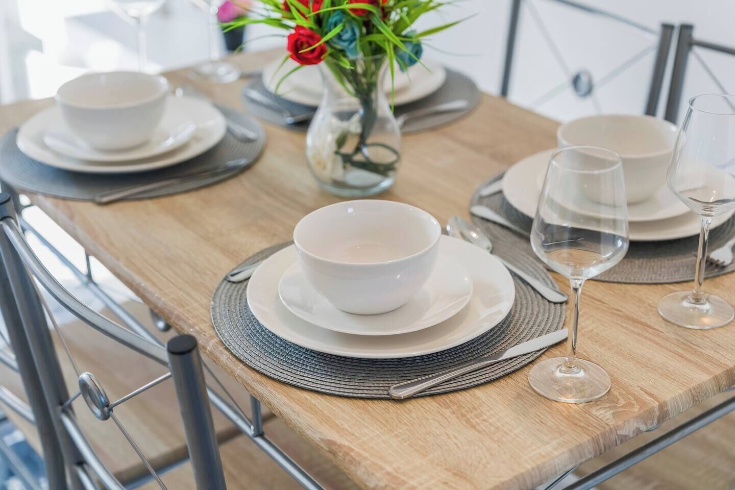 Una mesa de comedor dispuesta con platos, tazones y copas de vino, junto a una jarra con flores.