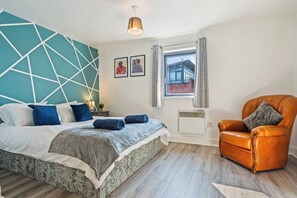 Quarto moderno com design geométrico na parede, cama com roupa de cama azul e branca, poltrona de couro e janela com vista.