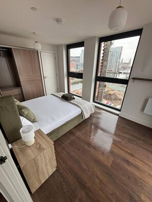 A modern bedroom with a large bed, a wooden wardrobe, and a view of a cityscape through large windows. - #132 Stylish Luxury Flat | Free Parking |City View (Merseyside)