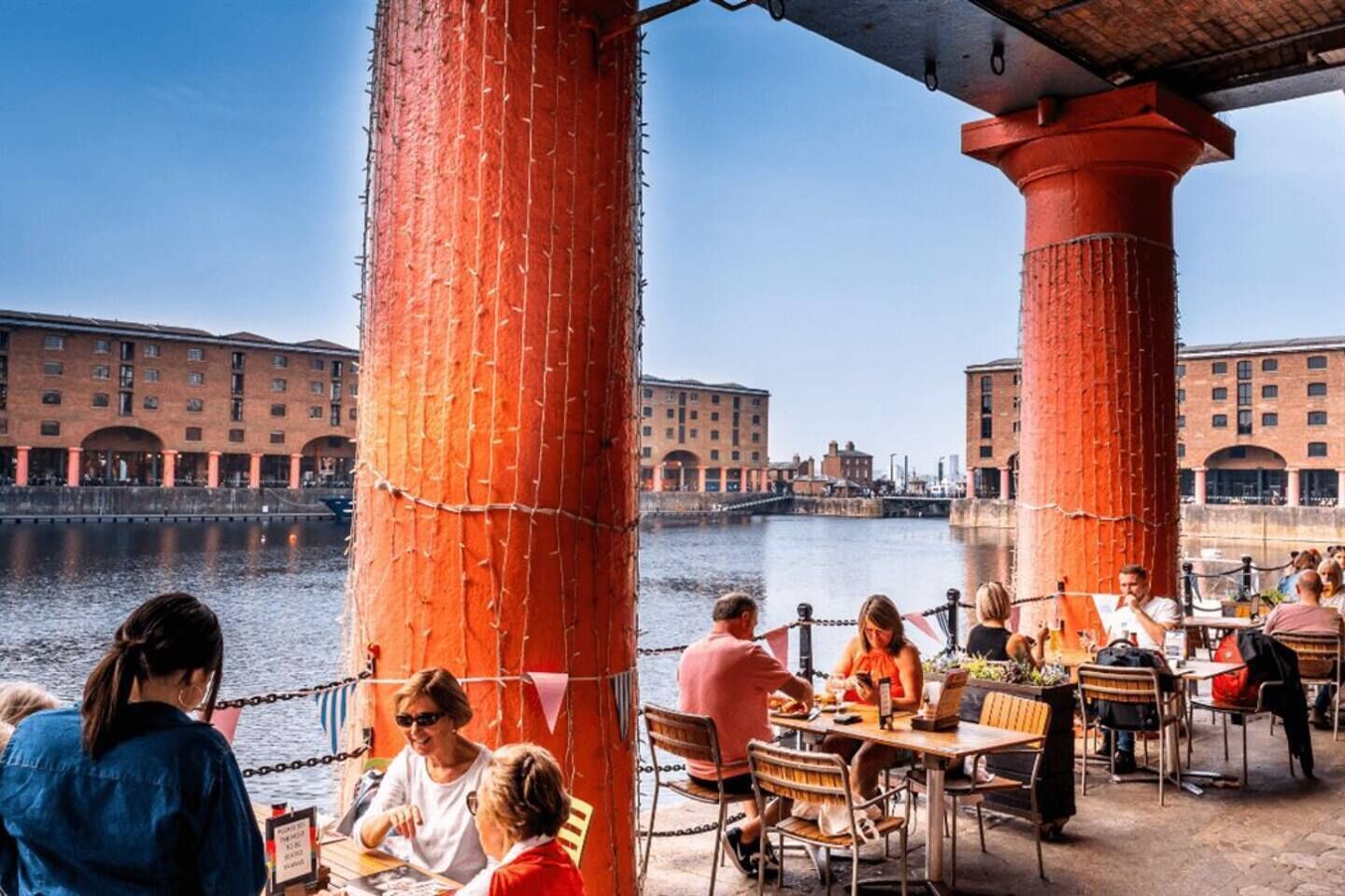 A waterfront restaurant with outdoor seating, red pillars, and a view of historic buildings across the water.