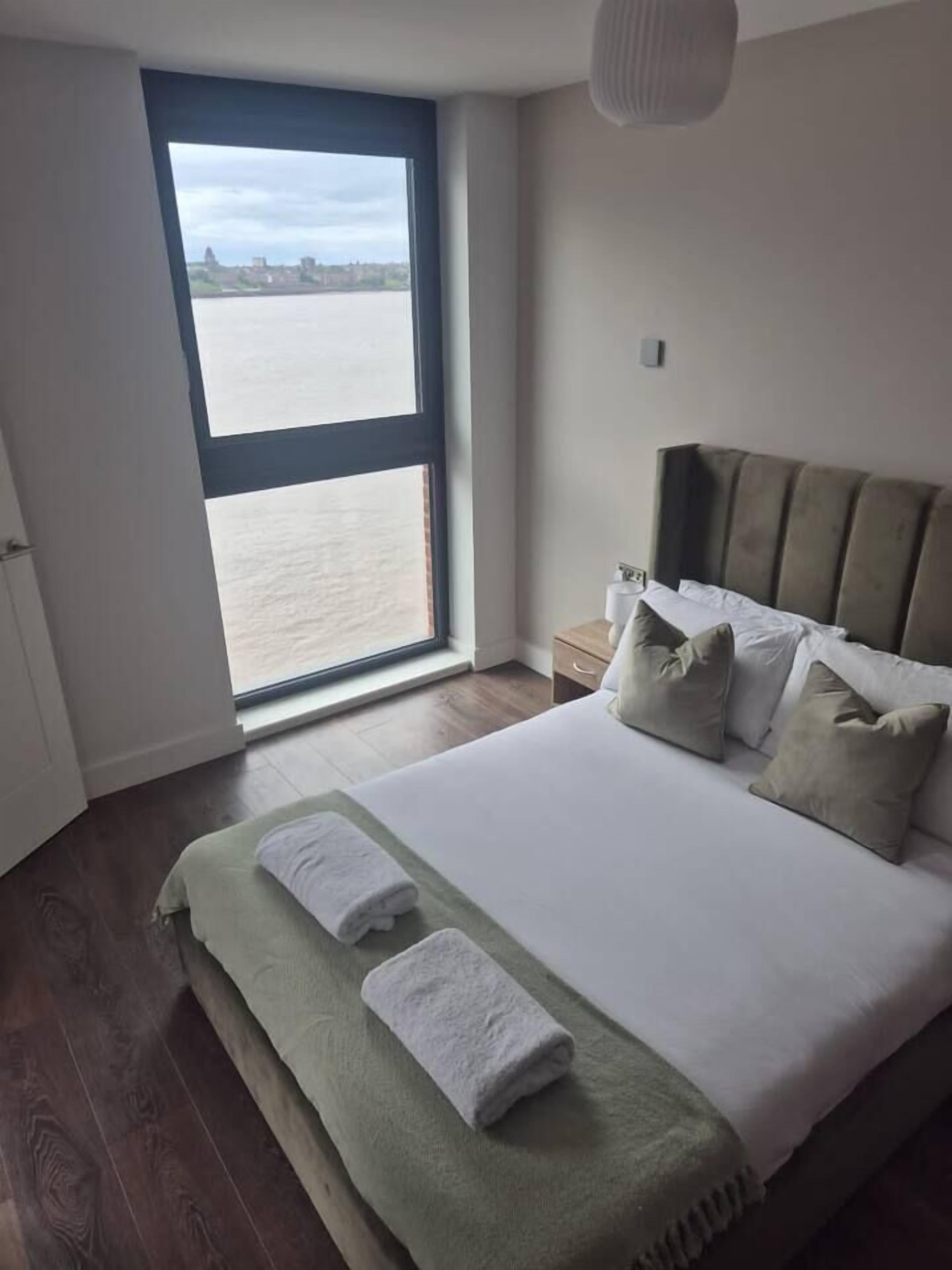 A bedroom with a large window, a bed with pillows, a bedside table, and a nightstand.
