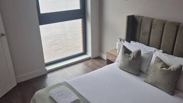 A bedroom with a large window, a bed with pillows, a bedside table, and a nightstand.