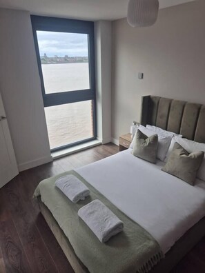 A bedroom with a large window, a bed with pillows, a bedside table, and a nightstand. - #132 Stylish Luxury Flat | Free Parking |City View (Merseyside)