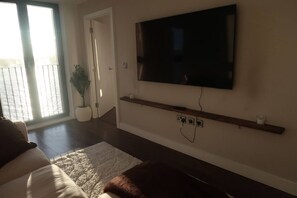 Ein Wohnzimmer mit einem an der Wand montierten Flachbildfernseher, einem Holzregal darunter und einem großen Fenster mit Blick ins Freie.
