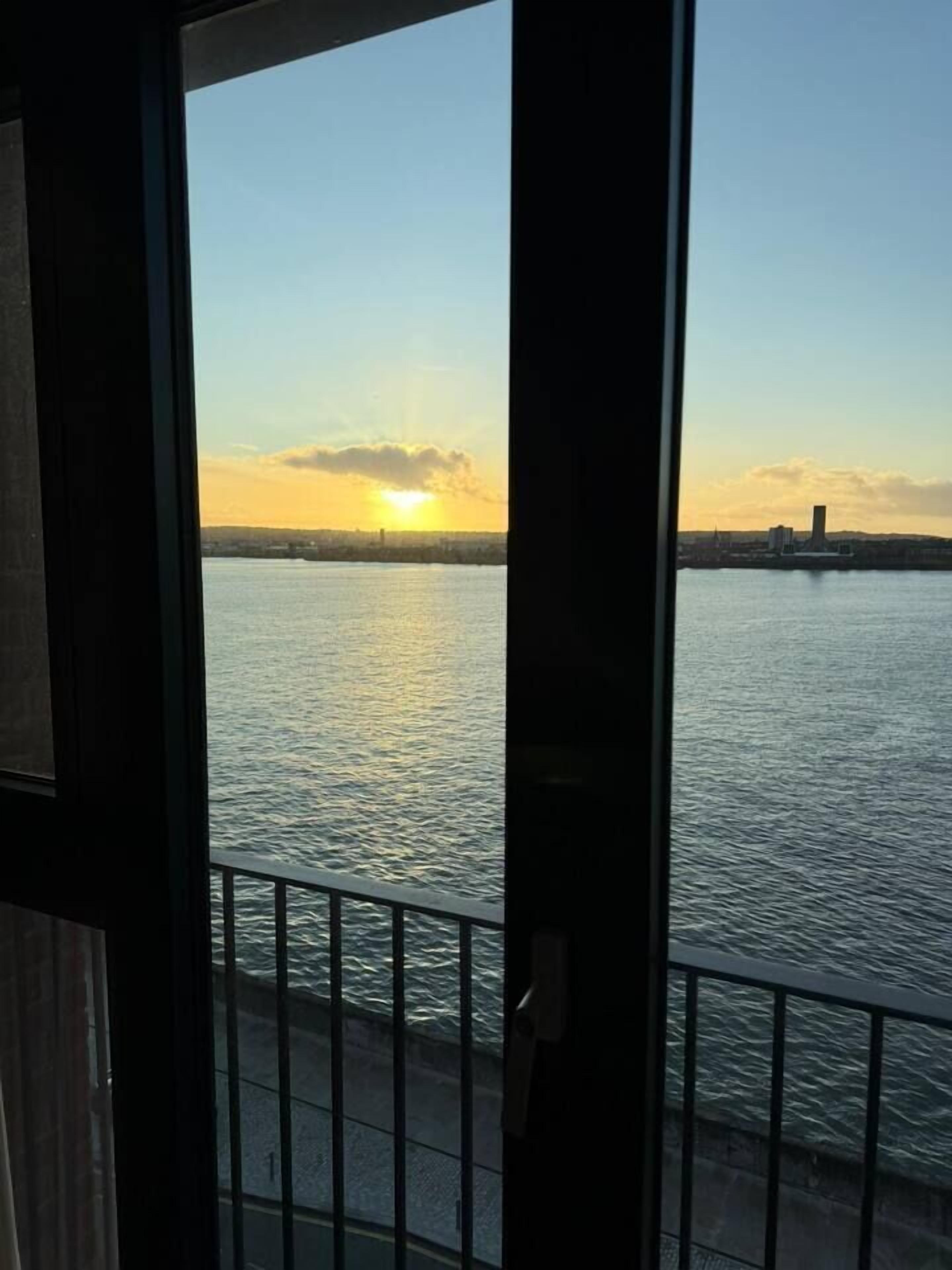A view of a body of water with a sunset, a distant city skyline, and a metal railing.