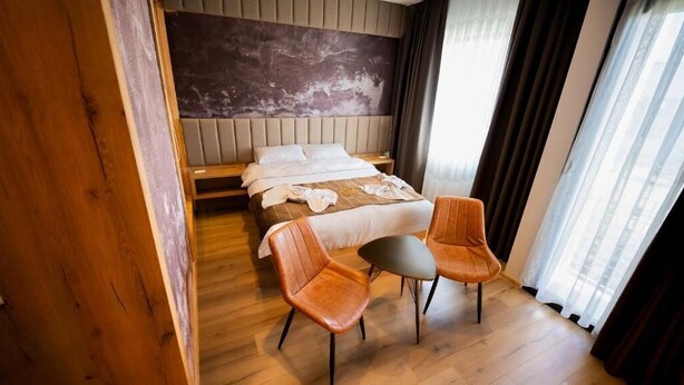 A hotel room with a bed, two chairs, a small table, and a view of the city through the window. - Ava Hotel Prishtina (Lipjan)