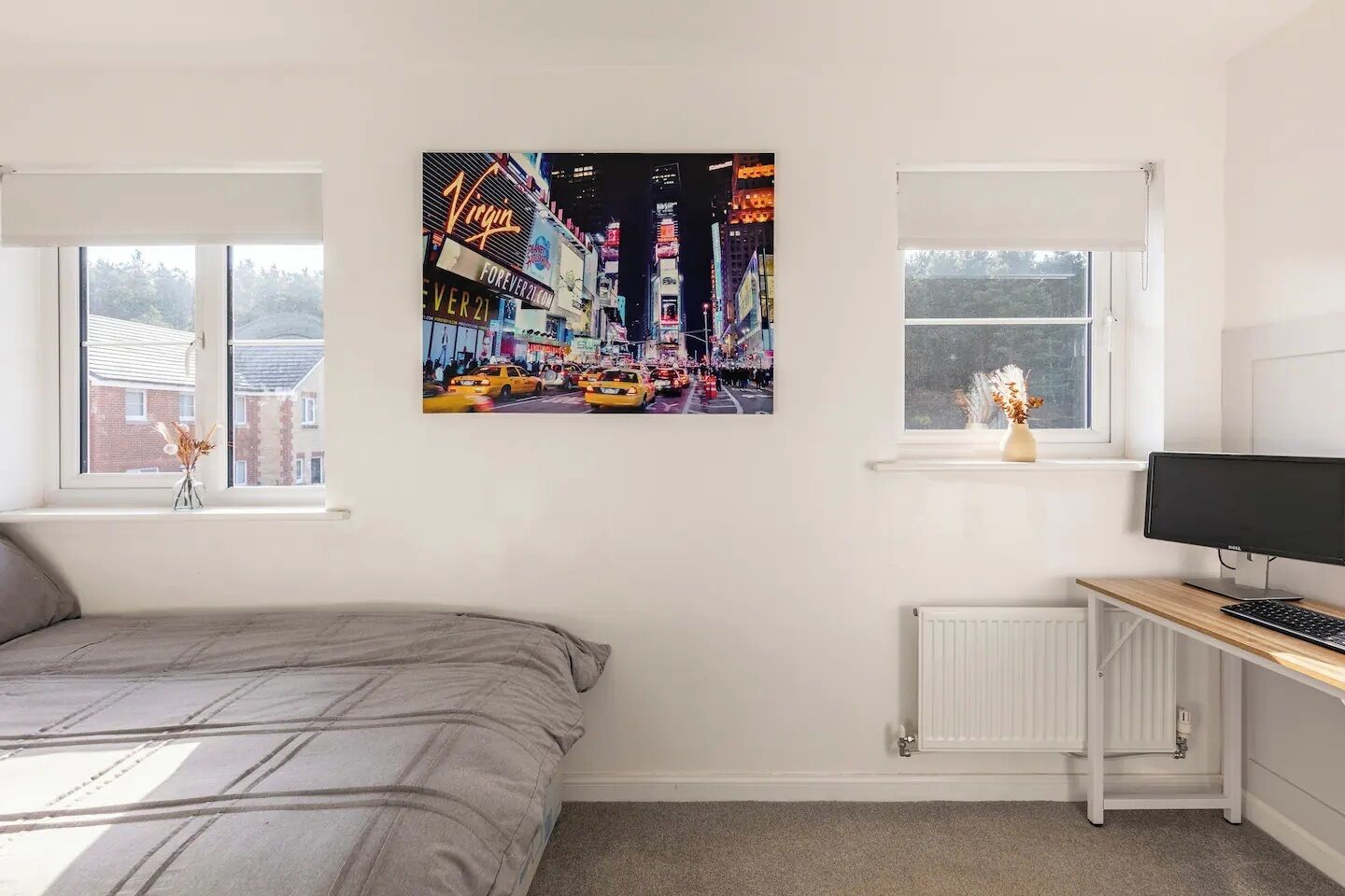 Une chambre à coucher avec un lit, un bureau avec un ordinateur, deux fenêtres et un tableau représentant un paysage urbain.