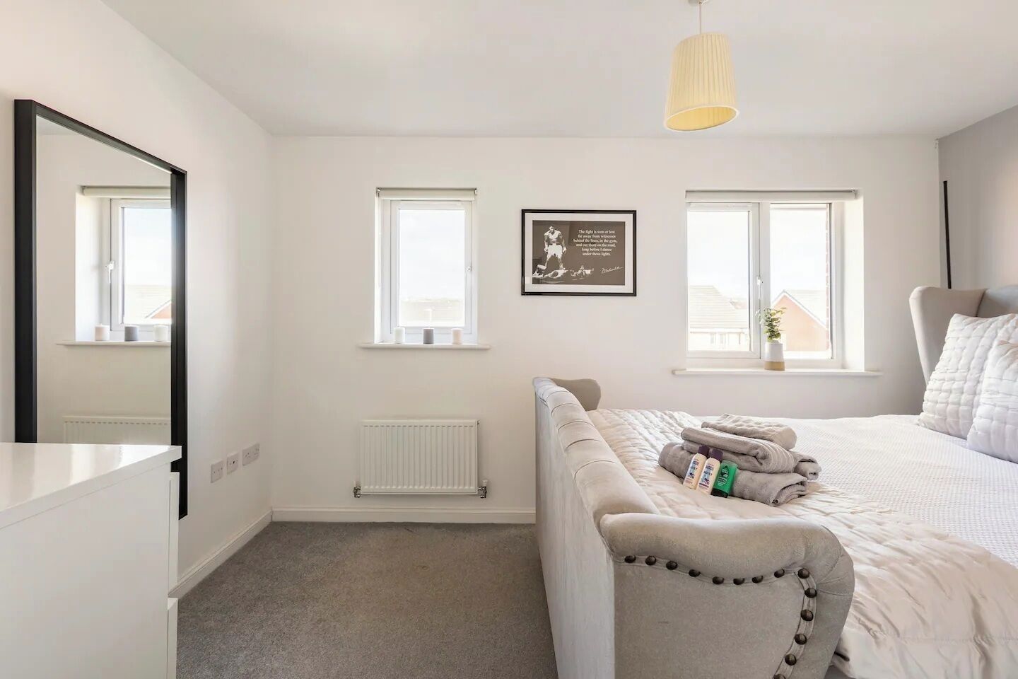 Une chambre à coucher comprenant un lit, une table de chevet, un radiateur, deux fenêtres et un luminaire suspendu.