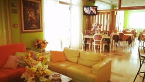 Un vestíbulo de hotel con un sofá amarillo, una zona de comedor con mesas y sillas, y un televisor montado en la pared.
