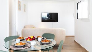 Een moderne woonkamer met een glazen eettafel, groene stoelen, een beige bank en een flatscreen televisie aan de muur.